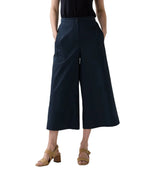 Model in Ann Mashburn's Drew Pant, wide-leg and cropped poplin style, paired with a black blouson top and tan heeled sandals, stands with hands in pockets against a white background.