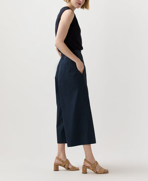 A person stands sideways in a blouson-style black sleeveless top, tan heeled sandals, and Ann Mashburn's Drew Pant—dark wide-leg cropped poplin trousers—set against a plain background.