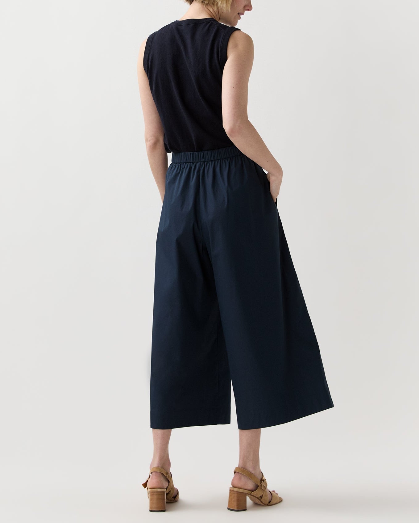 A person faces away, wearing a sleeveless black blouson top, Ann Mashburn Drew Pant in navy poplin, and tan heeled sandals against a plain light background.