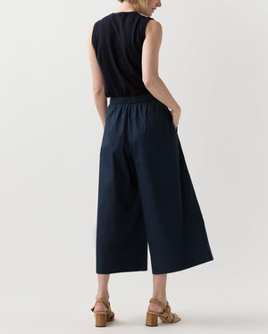A person faces away, wearing a sleeveless black blouson top, Ann Mashburn Drew Pant in navy poplin, and tan heeled sandals against a plain light background.