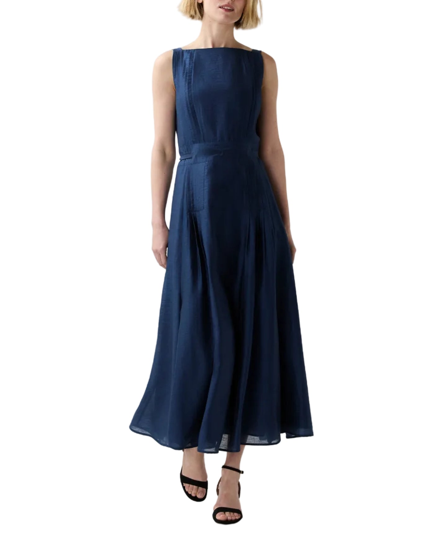 A woman wears the Ann Mashburn Rose Dress—a sleeveless, navy, ankle-length pleated dress with a fitted waist and square neckline—paired with black high-heeled sandals for chic evening style.