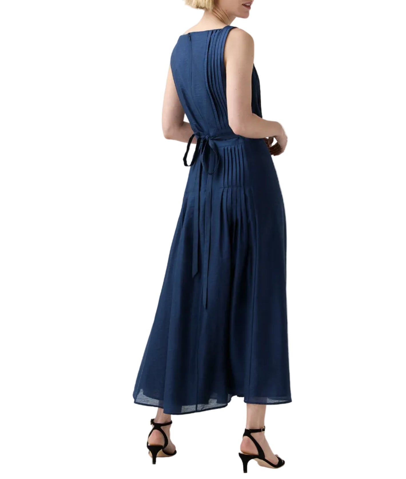 A woman wears the Ann Mashburn Rose Dress—an ankle-length, sleeveless navy pleated dress with a square neckline and tie-back—paired with black heeled sandals, ideal for elegant evening occasions.
