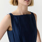 A person with short blonde hair wears the Ann Mashburn Rose Dress—a sleeveless navy blue pleated dress with detailing near the square neckline—standing against a plain light background.