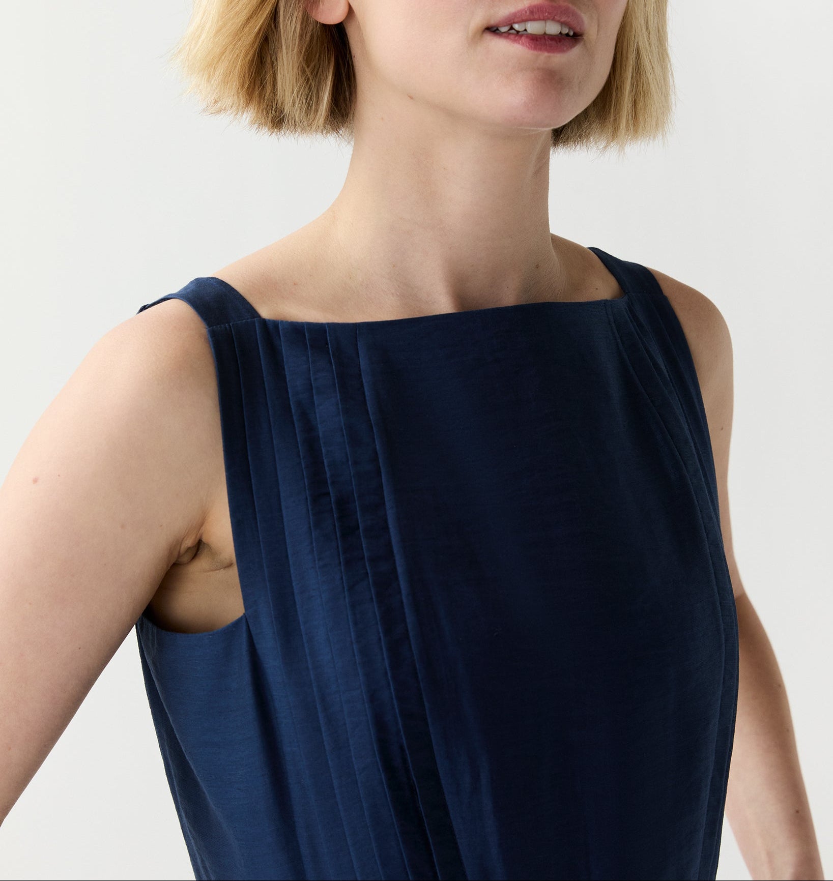 A person with short blonde hair wears the Ann Mashburn Rose Dress—a sleeveless navy blue pleated dress with detailing near the square neckline—standing against a plain light background.