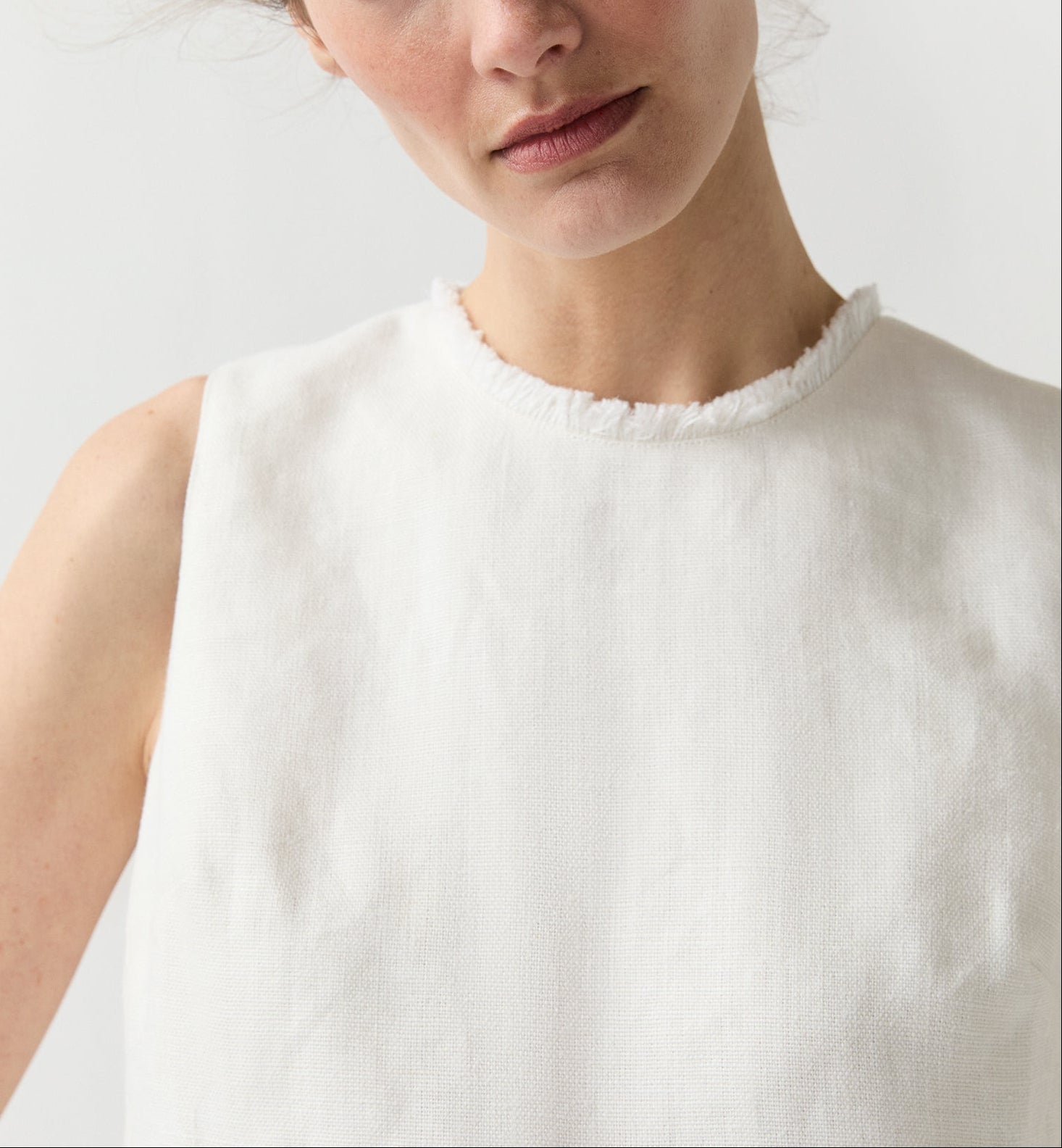 A person is shown from the shoulders up against a plain background, wearing the Ann Mashburn Mimi Linen Top—a sleeveless white Belgian linen shell with a frayed neckline.