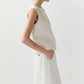 A woman in the Ann Mashburn Mimi Linen Top stands in profile with one hand in her pocket against a plain light background.