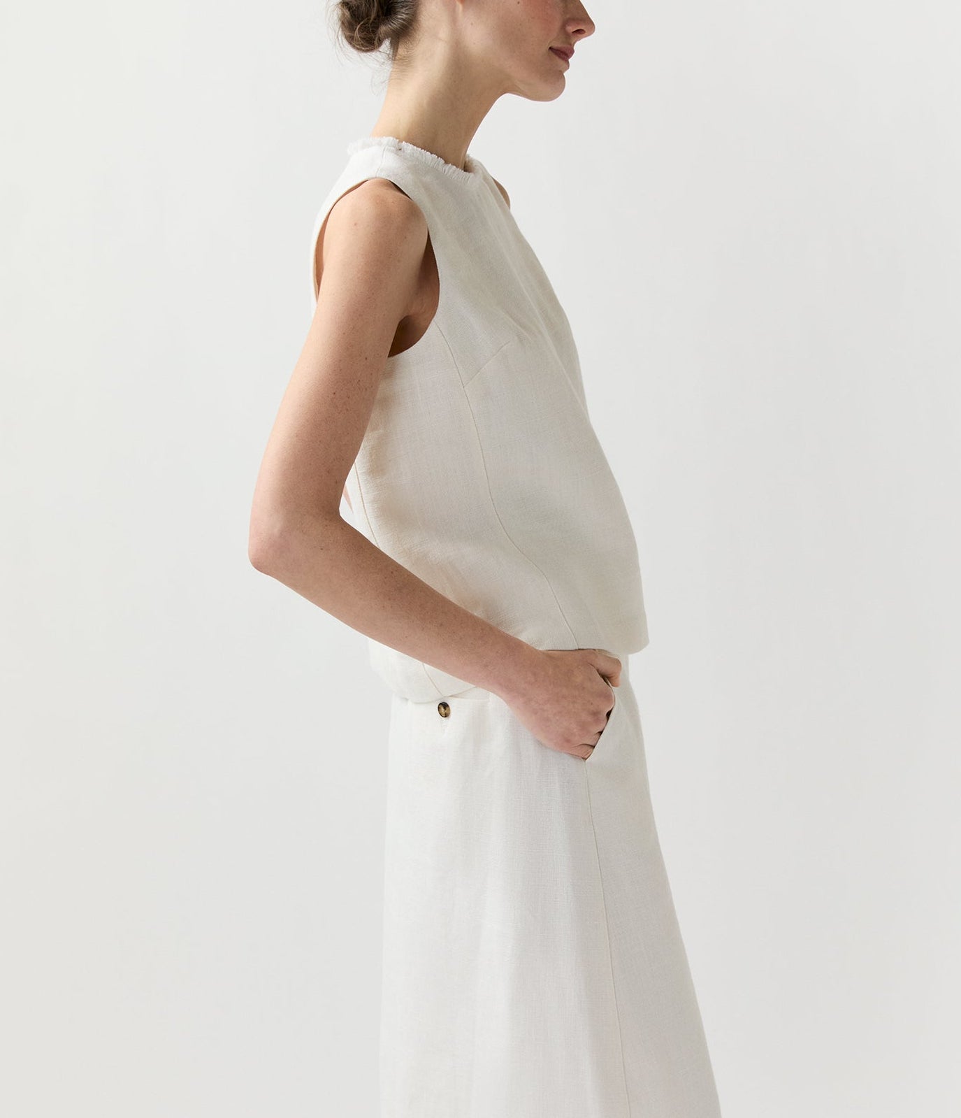 A woman in the Ann Mashburn Mimi Linen Top stands in profile with one hand in her pocket against a plain light background.