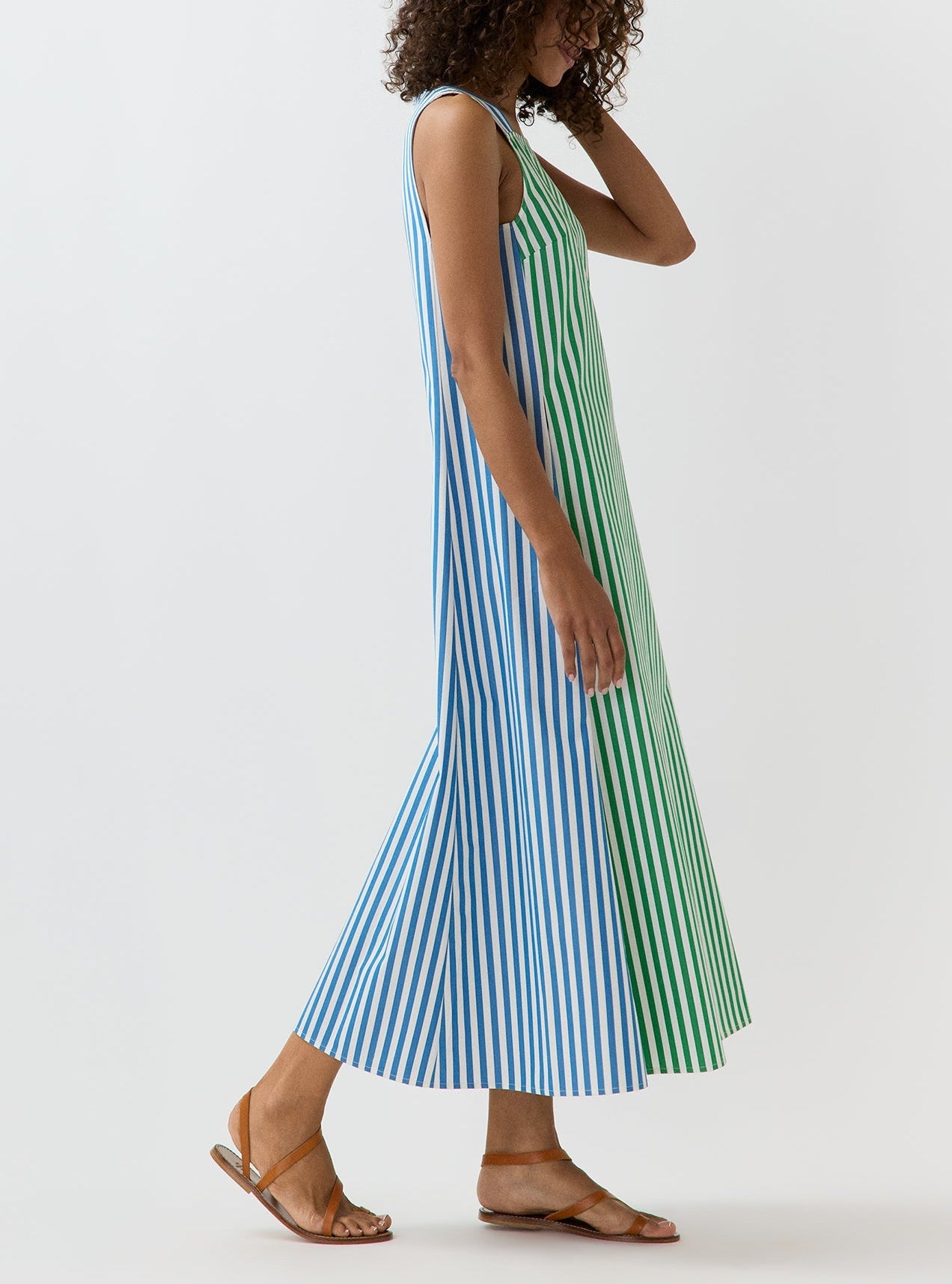 A woman wearing the Lise Maxi Dress by Ann Mashburn—a sleeveless, ankle-length A-line dress in striped Italian poplin with blue and green vertical stripes and a square neckline—pairs it with brown sandals against a plain white background.