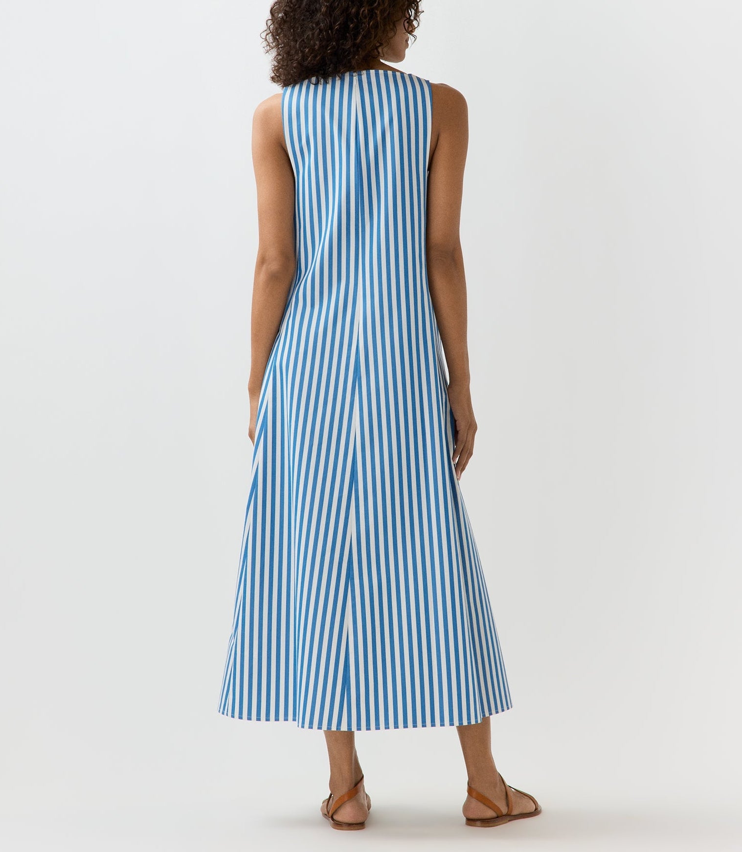 A woman faces away, wearing the Ann Mashburn Lise Maxi Dress—a sleeveless, ankle-length, A-line style in striped Italian poplin—with flat sandals, set against a plain light background.