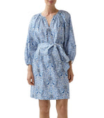 A person wearing the Ann Mashburn Genevieve Liberty Print Dress—a knee-length, blue and white romantic dress with a split neckline, long sleeves, and belted waist—stands against a plain background.