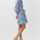 A woman in the Ann Mashburn Genevieve Liberty Print Dress, featuring a blue and white pattern and split neckline, pairs it with gold strappy heels against a plain white background.