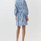 A woman, seen from behind, wears the Ann Mashburn Genevieve Liberty Print Dress with a blue and white pattern, split neckline, long sleeves, and beige heeled sandals.