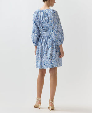 A woman, seen from behind, wears the Ann Mashburn Genevieve Liberty Print Dress with a blue and white pattern, split neckline, long sleeves, and beige heeled sandals.