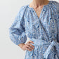 A woman wearing the Ann Mashburn Genevieve Liberty Print Dress, featuring a blue and white pattern, split neckline, long sleeves, and a matching trapunto-stitched belt at the waist stands with one hand on her hip.