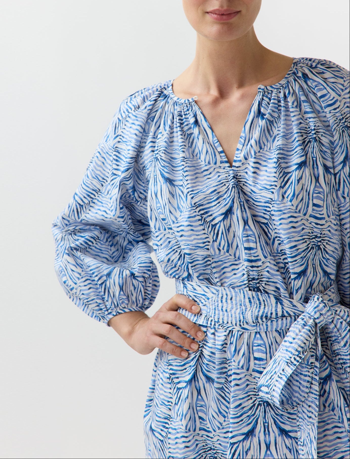 A woman wearing the Ann Mashburn Genevieve Liberty Print Dress, featuring a blue and white pattern, split neckline, long sleeves, and a matching trapunto-stitched belt at the waist stands with one hand on her hip.