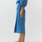 A person stands sideways wearing the Ann Mashburn Morgan Variegated Stripe Skirt, featuring blue and white vertical stripes, styled like a summer skirt, paired with black pointed-toe shoes.