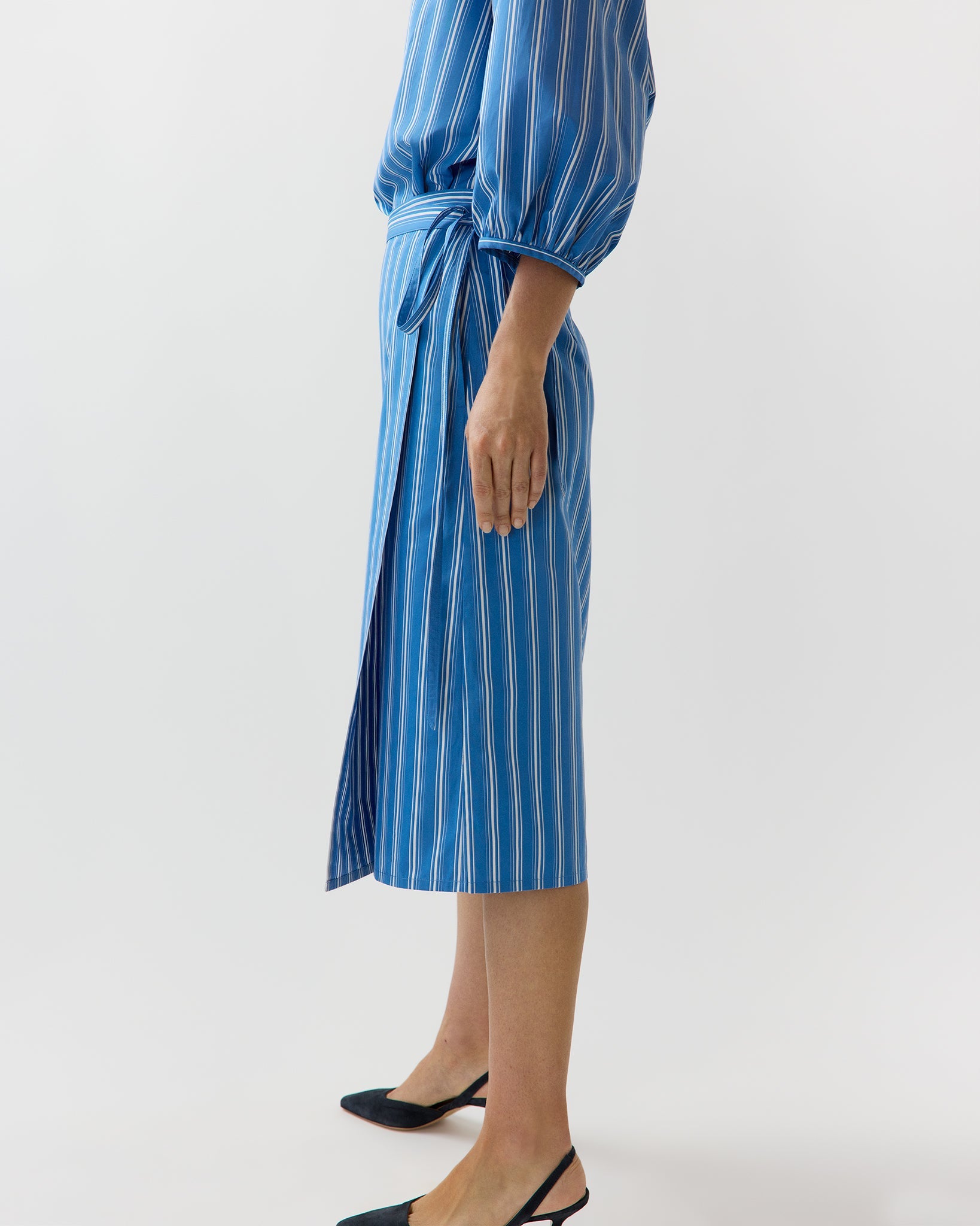 A person stands sideways wearing the Ann Mashburn Morgan Variegated Stripe Skirt, featuring blue and white vertical stripes, styled like a summer skirt, paired with black pointed-toe shoes.