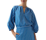 Model wearing the Ann Mashburn Victoria Variegated Stripe Blouse in blue and white Italian poplin, styled with a matching skirt, one hand in the skirt pocket, photographed against a white background.