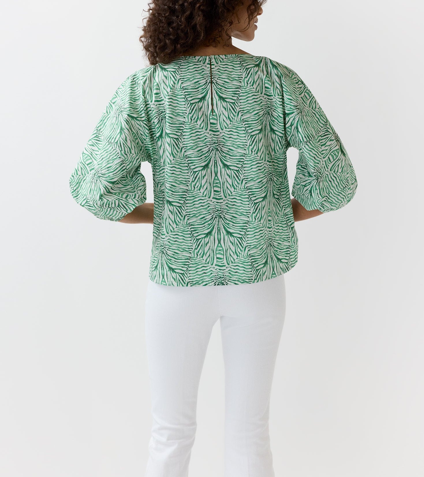 A person stands facing away, wearing the Elora Volume Liberty Top by Ann Mashburn—featuring a green and white zebra print with loose sleeves—and white pants, set against a plain white background.