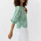 A woman wears the Ann Mashburn Elora Volume Liberty Top in green and white with three-quarter sleeves, paired with white pants against a plain backdrop—a breezy summer look.