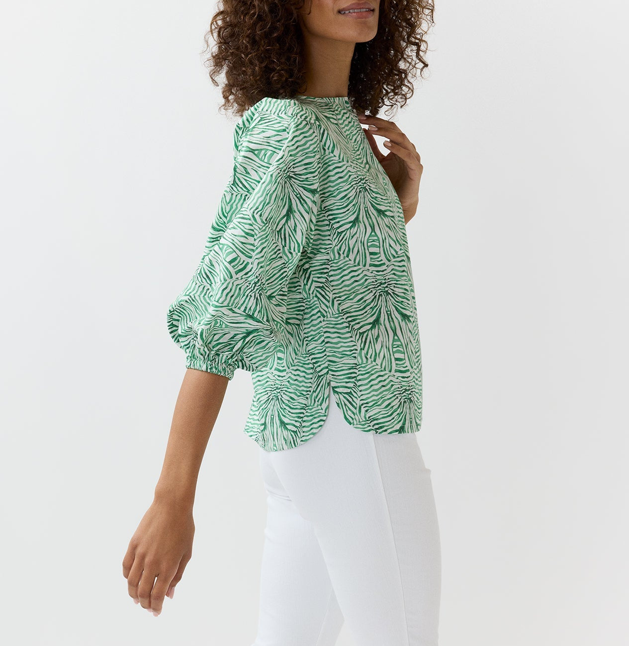A woman wears the Ann Mashburn Elora Volume Liberty Top in green and white with three-quarter sleeves, paired with white pants against a plain backdrop—a breezy summer look.