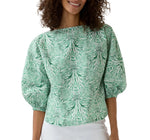 A woman wears the Elora Volume Liberty Top by Ann Mashburn—a green and white patterned blouse with puffed sleeves and a boat neckline—styled with a white skirt for an elegant summer look.