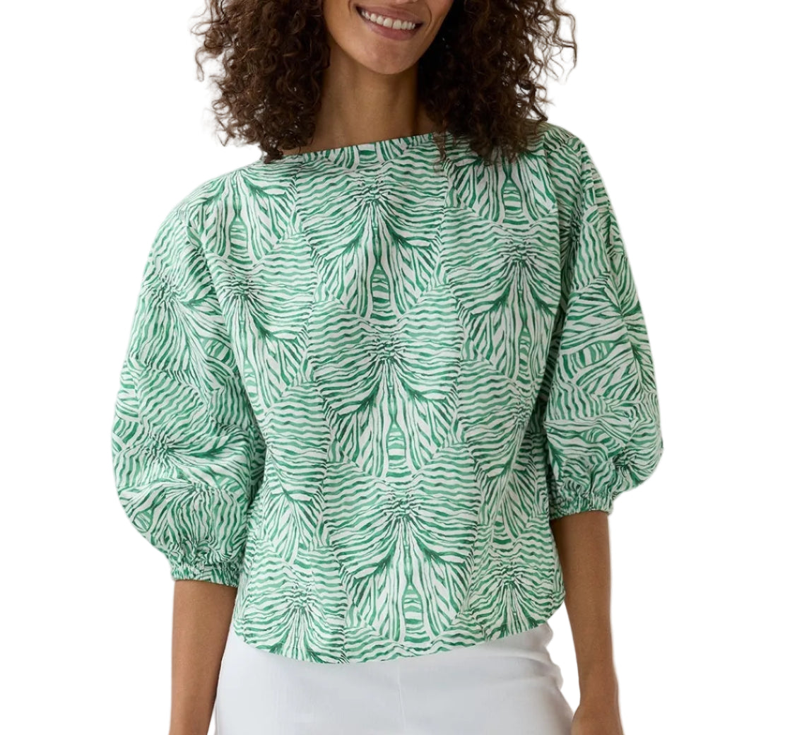 A woman wears the Elora Volume Liberty Top by Ann Mashburn—a green and white patterned blouse with puffed sleeves and a boat neckline—styled with a white skirt for an elegant summer look.