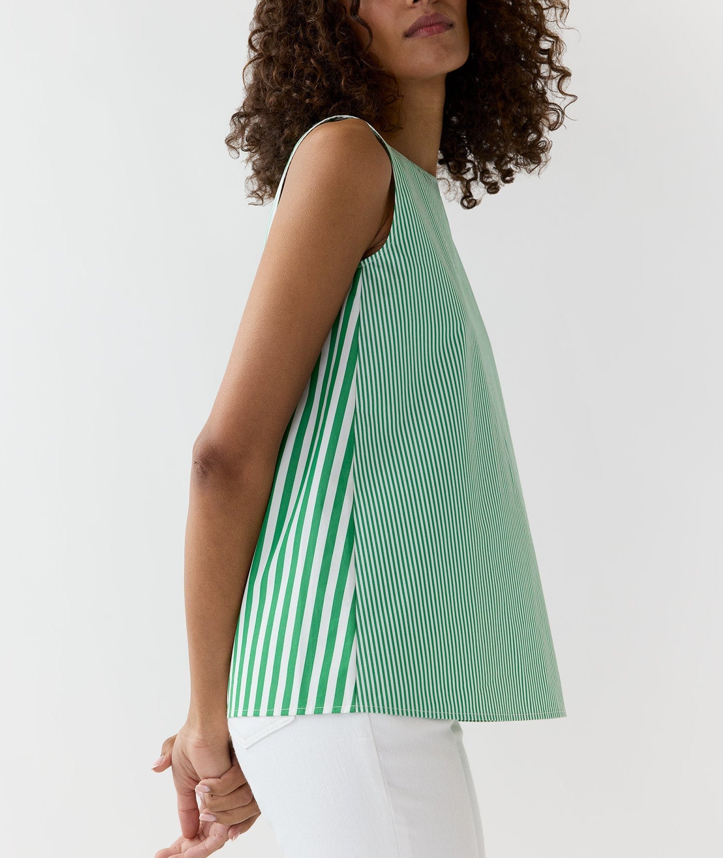 A woman wears the Ann Mashburn Bell Top—a simple tank in Italian striped poplin with green and white vertical stripes and a bateau neckline—paired with white pants, set against a plain white background.