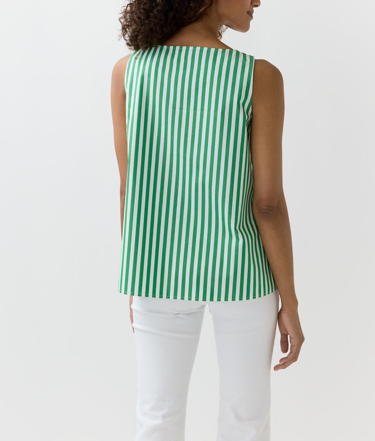 A woman is seen from the back wearing the Ann Mashburn Bell Top, a sleeveless green and white vertically striped Italian poplin, paired with white pants against a plain background.