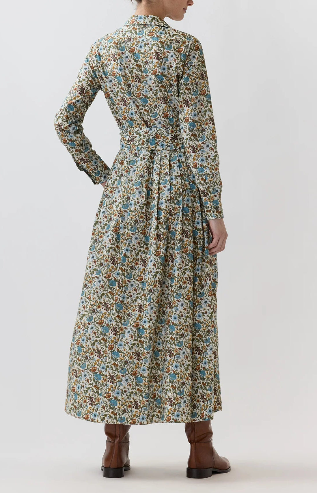A person stands with their back to the camera, wearing the Ann Mashburn Classic Shirtwaist Rachel Liberty Dress—an ankle-length, long-sleeve floral dress—paired with brown ankle boots against a plain light background.