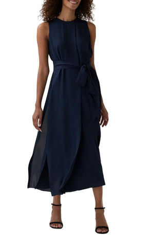 A woman wears the Ann Mashburn Scarlett Silk Georgette Dress—a sleeveless, navy blue dress with a belted waist and side slits—paired with black ankle-strap heels, standing against a white background.