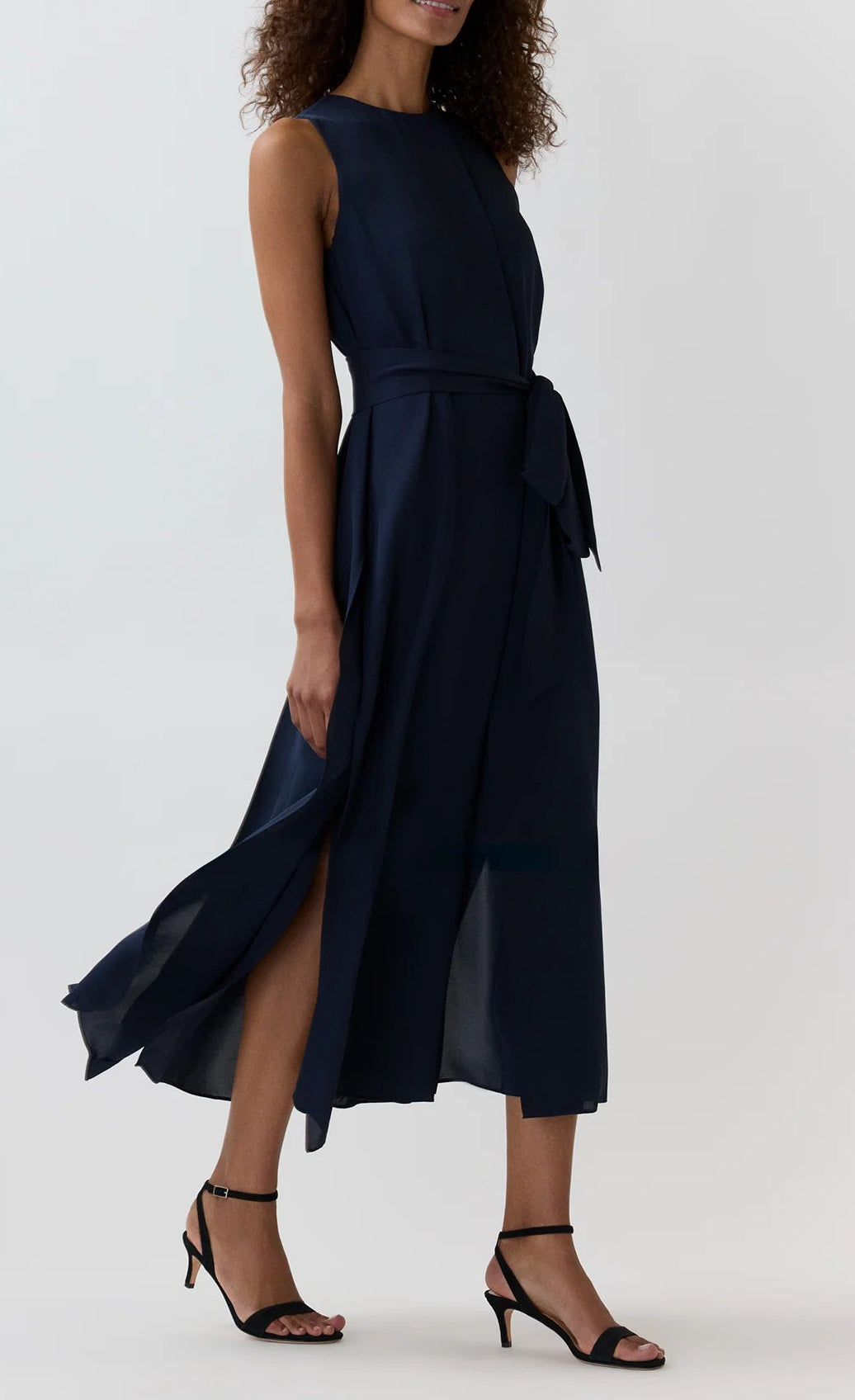 A woman is wearing the Ann Mashburn Scarlett Silk Georgette Dress—a sleeveless, navy blue midi dress with a waist tie—paired with black heeled sandals, standing against a plain light background. This lightweight dress from Ann Mashburn drapes beautifully, offering effortless style and elegance.