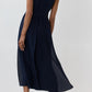 An elegant woman stands with her back to the camera, wearing the Ann Mashburn Scarlett Silk Georgette Dress—a sleeveless, dark navy midi style crafted from double silk georgette and cinched at the waist with a belt—paired with black heels, set against a plain light background.