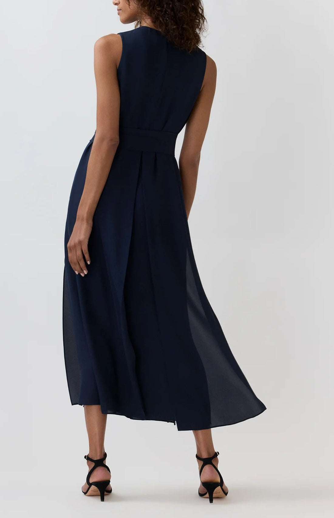 An elegant woman stands with her back to the camera, wearing the Ann Mashburn Scarlett Silk Georgette Dress—a sleeveless, dark navy midi style crafted from double silk georgette and cinched at the waist with a belt—paired with black heels, set against a plain light background.