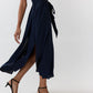 A person wears the Ann Mashburn Scarlett Silk Georgette Dress—a sleeveless navy dress featuring a belt and slit—paired with black heeled sandals, walking against a plain background.
