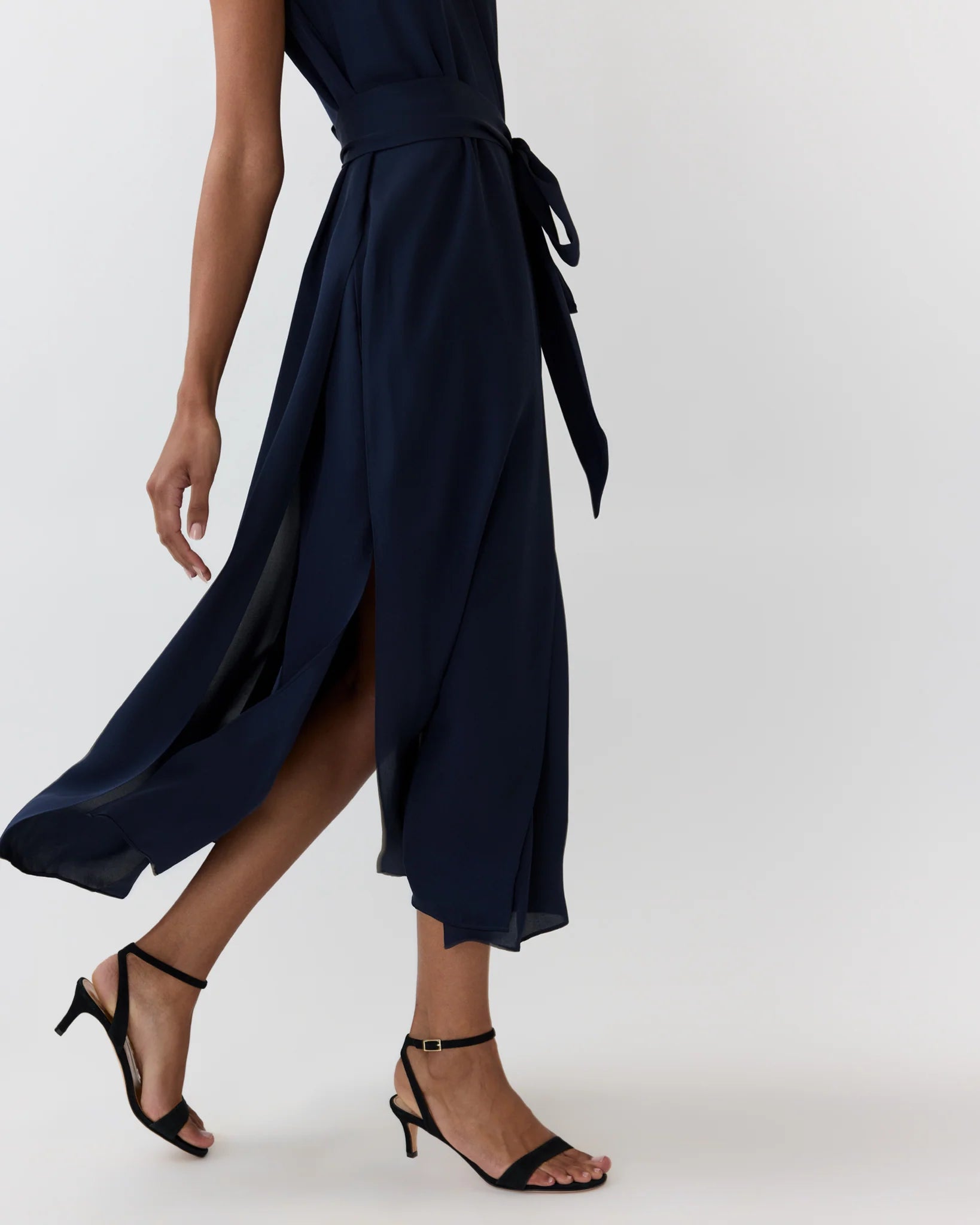 A person wears the Ann Mashburn Scarlett Silk Georgette Dress—a sleeveless navy dress featuring a belt and slit—paired with black heeled sandals, walking against a plain background.