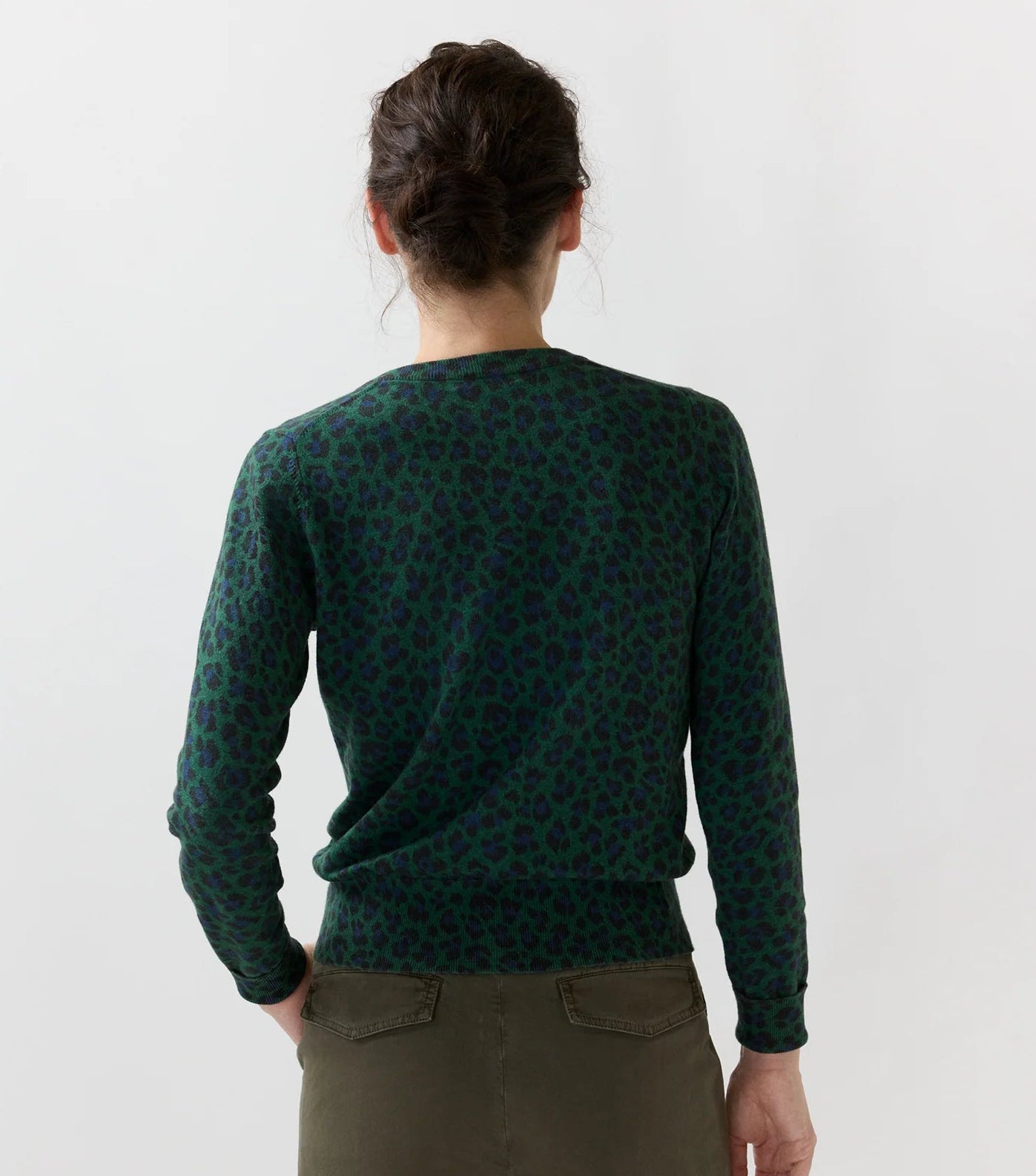 A person with dark hair in a bun, wearing the Ann Mashburn Rosie Cotton Silk Leopard Cardigan and olive pants, is shown from the back against a plain light background.
