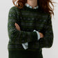 A person with long wavy hair stands with arms crossed against a plain background, wearing the Ann Mashburn Alpine Fair Isle Cashmere Silk Sweater in green over a collared shirt.