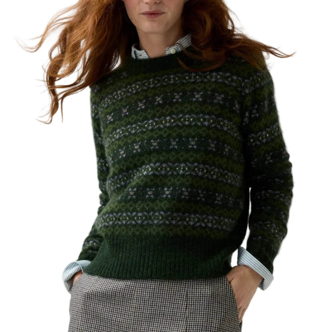 A woman with long, wavy reddish hair wears the Ann Mashburn Alpine Fair Isle Cashmere Silk Sweater in dark green over a striped collared shirt, paired with brown plaid pants. The background is plain white.