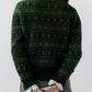 A person facing away, wearing the Ann Mashburn Alpine Fair Isle Cashmere Silk Sweater in dark green over a light blue collared shirt and plaid skirt, with hands clasped behind their back.
