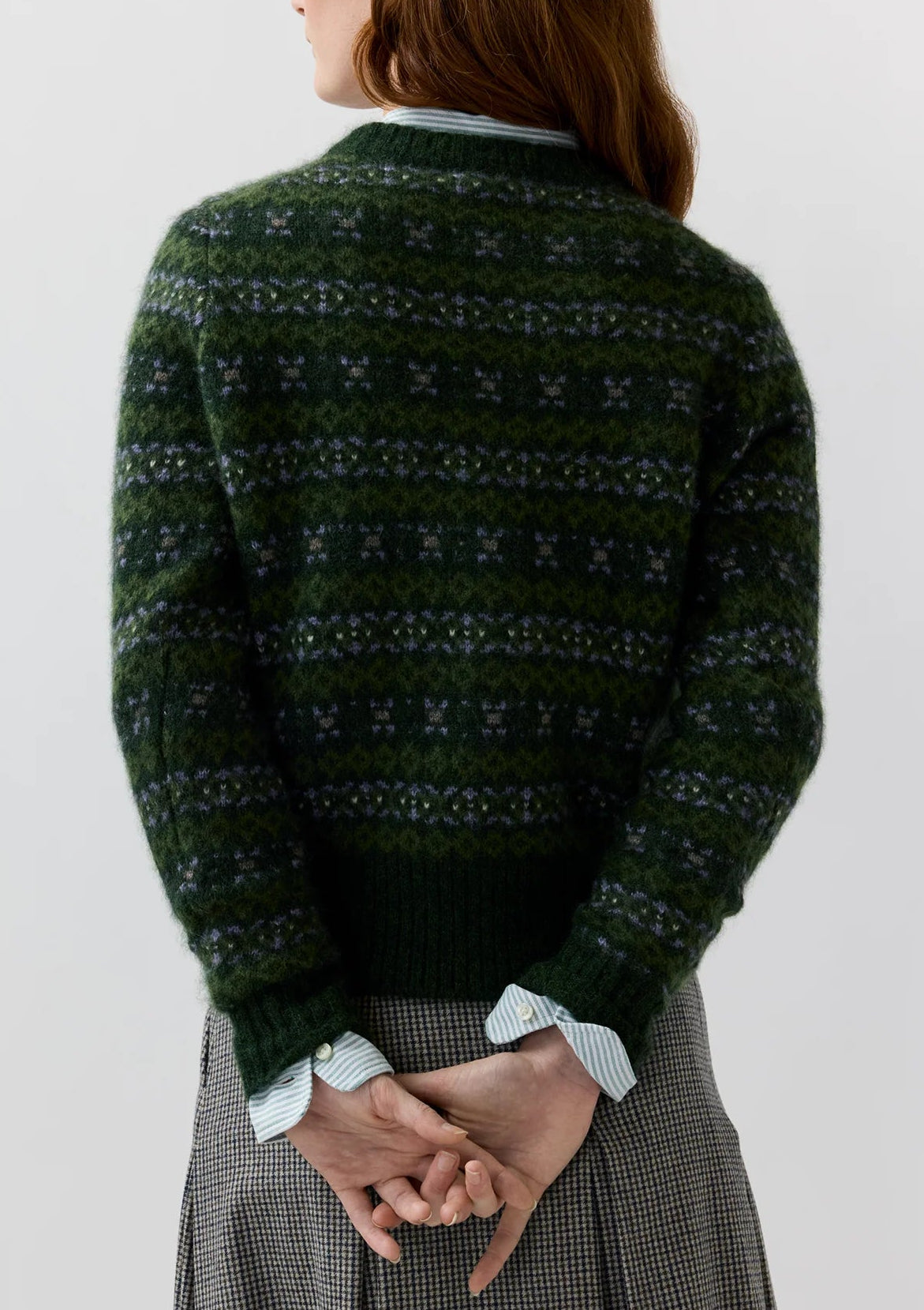 A person facing away, wearing the Ann Mashburn Alpine Fair Isle Cashmere Silk Sweater in dark green over a light blue collared shirt and plaid skirt, with hands clasped behind their back.