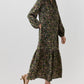 A person wearing an Aba Silk Maxi Dress in a dark green floral print with a belt, long sleeves, tiered skirt, and elasticated cuffs, styled with brown pointed-toe slingback heels.