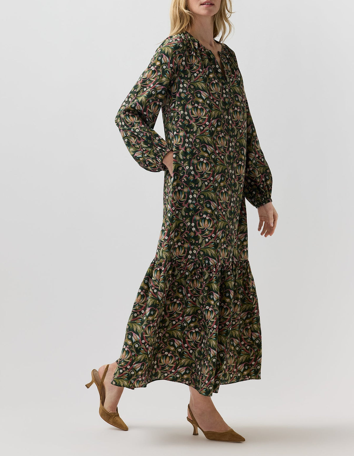A person wearing an Aba Silk Maxi Dress in a dark green floral print with a belt, long sleeves, tiered skirt, and elasticated cuffs, styled with brown pointed-toe slingback heels.
