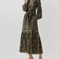 A dark green botanical print Aba Silk Maxi Dress featuring long sleeves, tiered skirt, elasticated cuffs, and a tie waist, worn with brown suede pumps.