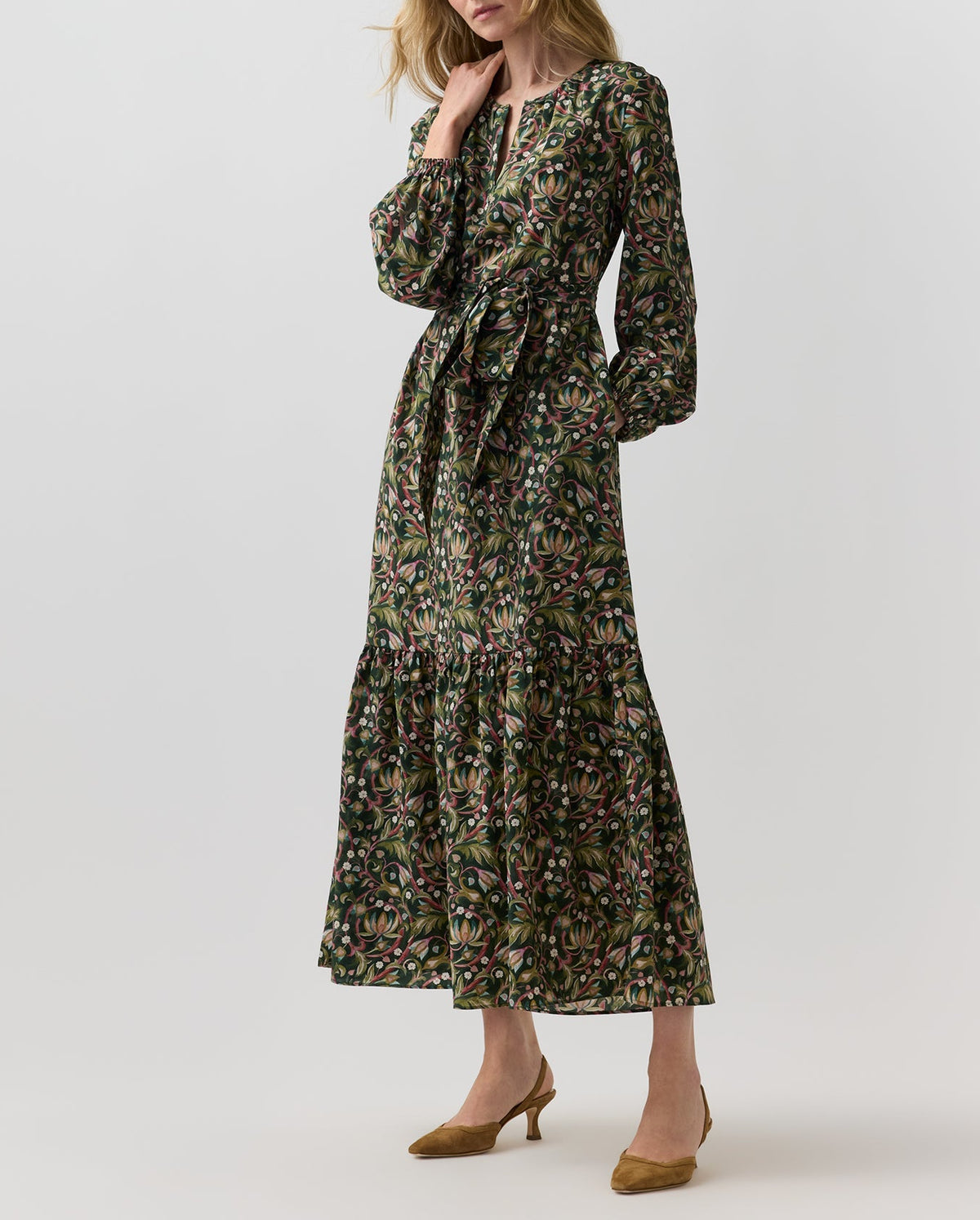 A dark green botanical print Aba Silk Maxi Dress featuring long sleeves, tiered skirt, elasticated cuffs, and a tie waist, worn with brown suede pumps.