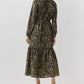 A woman seen from behind wearing the dark green floral Aba Silk Maxi Dress, featuring a tiered skirt, elasticated cuffs, and a belt styled with tan heels for a party silhouette.