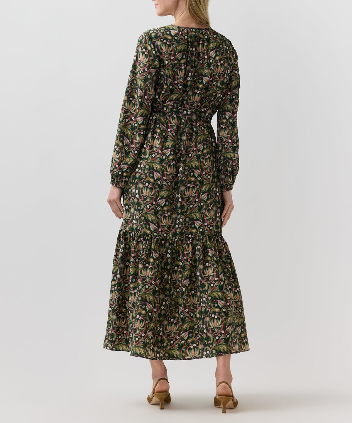 A woman seen from behind wearing the dark green floral Aba Silk Maxi Dress, featuring a tiered skirt, elasticated cuffs, and a belt styled with tan heels for a party silhouette.