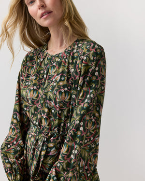 The Ann Mashburn Aba Silk Maxi Dress features a dark green botanical print with pink and white flowers, scrolling vines, a belt, and elasticated cuffs in a long-sleeved party silhouette.