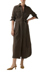 A person wearing the Ann Mashburn Maude Crinkle Cotton/Wool Dress—a long-sleeve, belted brown midi shirt dress with a collar and pockets made from textured fabric—stands with one hand on their hip and pairs the look with tan heeled shoes.