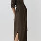 A woman stands against a plain light background wearing the Ann Mashburn Maude Crinkle Cotton/Wool Dress—a long-sleeved, belted brown shirt dress featuring a textured finish, front pockets, and side slits—paired with tan slingback heels.